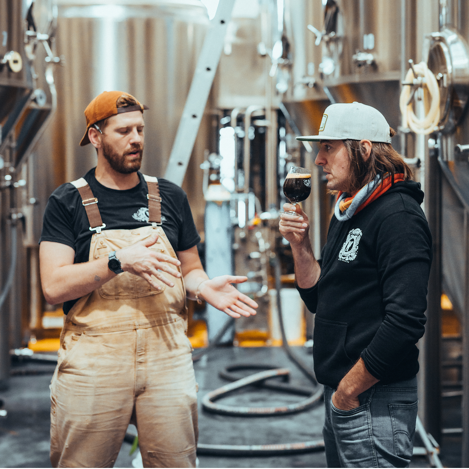 Two men in a brewery, one holding a glass of wine, with brewing equipment in the background.