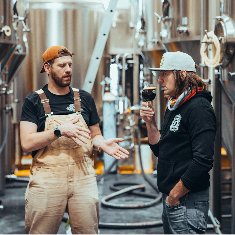 Two men in a brewery, one holding a glass of wine, with brewing equipment in the background.